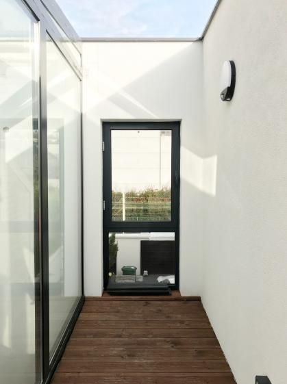 Cette image montre une maison moderne à façade blanche avec des fenêtres étroites et verticales. L'entrée dispose d'une porte gris foncé entourée de luminaires muraux. À gauche, un mur en béton texturé borde une allée où sont garés des vélos. À droite, un jardin bien entretenu avec un arbre aux feuilles rouges apporte une touche naturelle. L'ensemble dégage une atmosphère minimaliste et épurée.