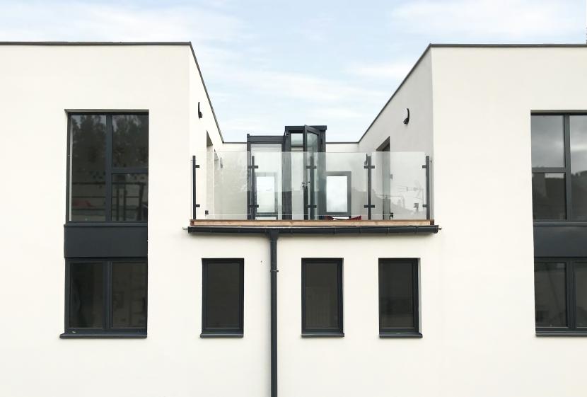 Cette image montre une maison moderne à façade blanche avec des fenêtres étroites et verticales. L'entrée dispose d'une porte gris foncé entourée de luminaires muraux. À gauche, un mur en béton texturé borde une allée où sont garés des vélos. À droite, un jardin bien entretenu avec un arbre aux feuilles rouges apporte une touche naturelle. L'ensemble dégage une atmosphère minimaliste et épurée.