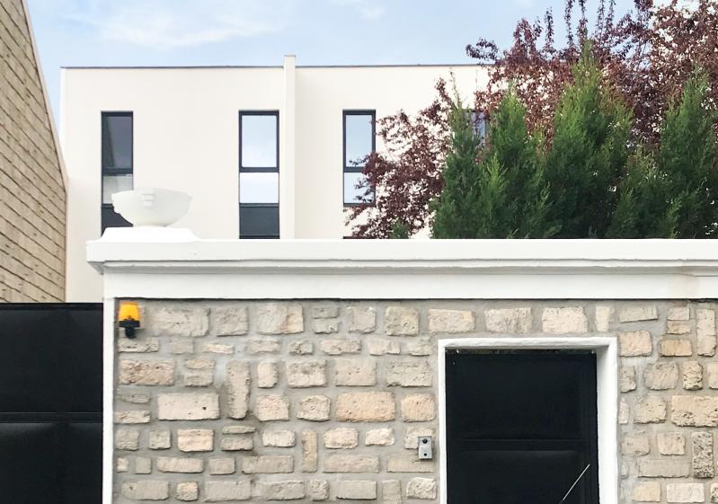 Cette image montre une maison moderne à façade blanche avec des fenêtres étroites et verticales. L'entrée dispose d'une porte gris foncé entourée de luminaires muraux. À gauche, un mur en béton texturé borde une allée où sont garés des vélos. À droite, un jardin bien entretenu avec un arbre aux feuilles rouges apporte une touche naturelle. L'ensemble dégage une atmosphère minimaliste et épurée.