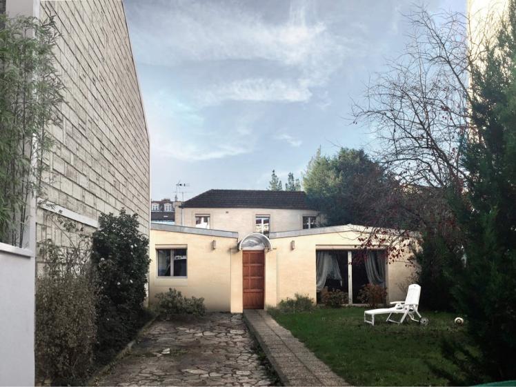 Cette image montre une maison moderne à façade blanche avec des fenêtres étroites et verticales. L'entrée dispose d'une porte gris foncé entourée de luminaires muraux. À gauche, un mur en béton texturé borde une allée où sont garés des vélos. À droite, un jardin bien entretenu avec un arbre aux feuilles rouges apporte une touche naturelle. L'ensemble dégage une atmosphère minimaliste et épurée.