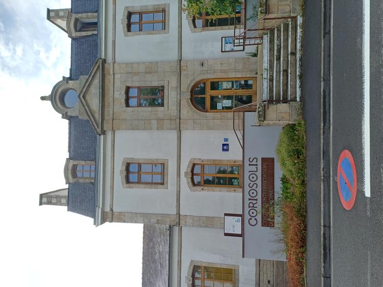Façade principale : un musée qui prend place dans l'ancienne école du village de Corseul. Fronton pierre & ordonnancement des ouvertures. Passerelle qui serpente dans la végétation et accompagne les visiteurs jusqu'à l'entrée du musée.