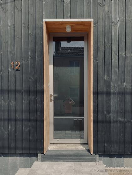 Façade avec porte entrée Façade en bardage noir, isolation en laine de bois, menuiserie aluminium et tableau en Douglas.