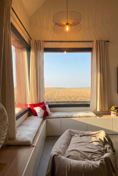 Cet espace du séjour est aménagé comme un coin calme et confortable, ouvert sur le paysage par une large baie vitrée. L’assise intégrée sous la fenêtre crée un lieu agréable pour lire ou se détendre tout en renforçant la relation entre l’espace de vie et l’extérieur.