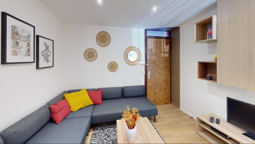 Séjour lumineux avec canapé d’angle gris, coussins colorés, table basse en bois, meuble TV et rangements muraux, dans un appartement rénové en colocation