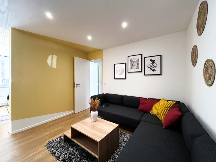 Salon de colocation avec canapé d’angle, mur jaune décoratif, éclairage encastré et accès vers cuisine, créant un espace convivial et chaleureux