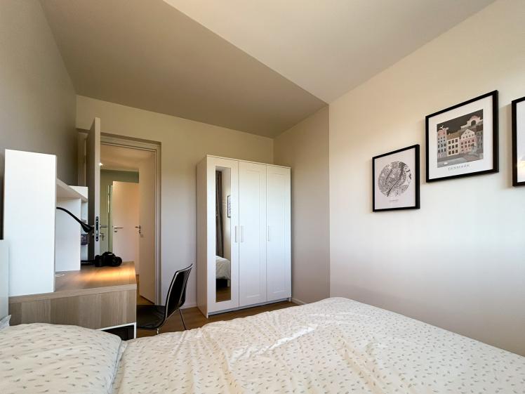 Chambre de colocation avec lit double, bureau en bois, armoire avec miroir et décoration murale, offrant un espace fonctionnel et lumineux