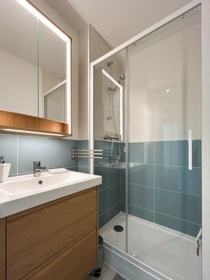Salle de bain avec douche vitrée, carrelage blanc et bleu, meuble vasque en bois et miroir éclairé, rénovée pour une colocation