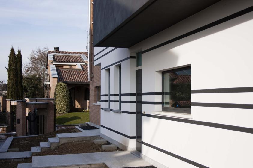 Détail façade – traitement des modénatures et insertion contextuelle Détail de façade latérale d'une maison jumelée contemporaine à Bellusco, Italie — enduit blanc avec bandeaux horizontaux sombres, menuiseries anthracite, insertion dans tissu pavillonnaire existant