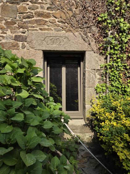 Une photographie en gros plan d'une petite fenêtre intégrée dans un vieux mur en pierres dorées et brunes. L'encadrement de la fenêtre est constitué de gros blocs de granit sculptés, et le linteau supérieur porte une date gravée à l'intérieur d'un cartouche géométrique (semblant indiquer 1724). La fenêtre moderne a des montants brun clair ou beige. Le mur est en partie recouvert de lierre grimpant sur la droite, tandis qu'un grand buisson d'hortensia aux feuilles bien vertes occupe le premier plan à gauche.