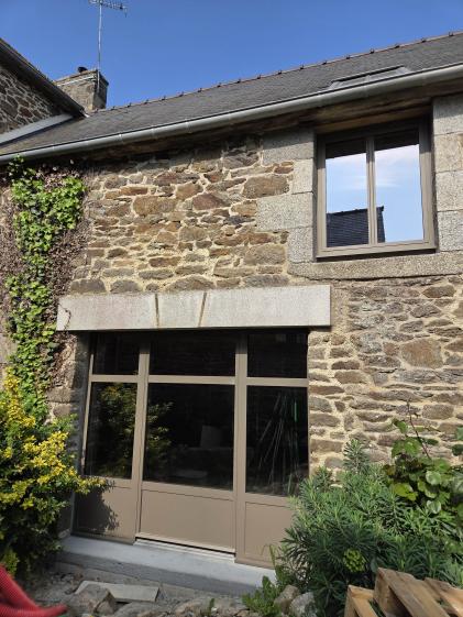Une photographie montrant une partie de la façade en rénovation d'une maison en pierre traditionnelle. Au rez-de-chaussée, une grande baie vitrée moderne à trois vantaux et soubassement plein (de couleur brun clair/taupe) est installée sous un imposant linteau en granit gris composé de trois blocs. À l'étage, juste au-dessus et décalée sur la droite, se trouve une fenêtre assortie encadrée de larges pierres de taille en granit, dans laquelle se reflète le ciel bleu.
