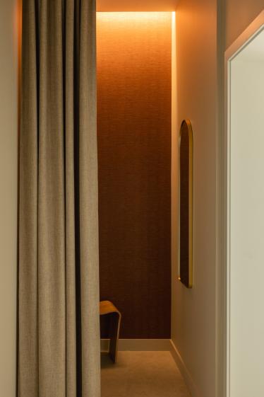 Cabine d'essayage au revêtement mural terracotta, éclairage LED indirect chaud, rideau en lin naturel et tabouret en bois, design spa luxe BEROA Arcachon par Eraiki StudioCopier