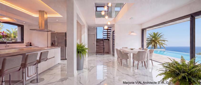 Vue du séjour de la maison contemporaine haut de gamme d'une surface plancher de 180 m². Grands volumes blancs lumineux, grandes ouvertures et lumière zénithale. Sol en marbre blanc, escalier métallique épuré, garde-corps en verre.