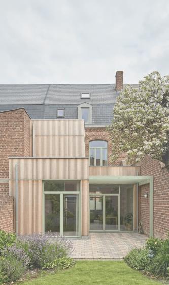 Perspective depuis le jardin Façade arrière en brique d'une maison ancienne, étendue par des volumes contemporains en bardage bois clair et menuiseries vert clair ouvrant sur un jardin planté.
