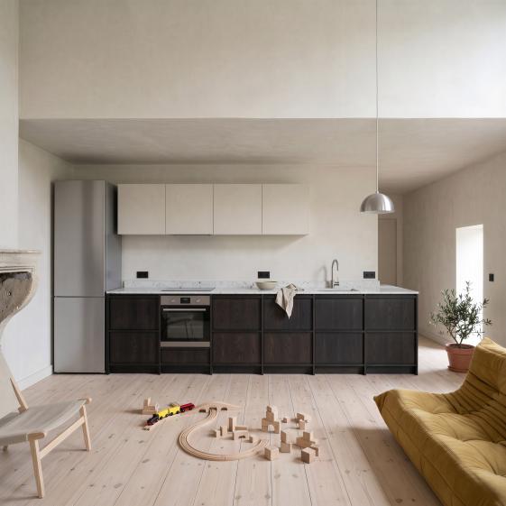 Un séjour-cuisine en réhabilitation, cadré frontalement : linéaire de cuisine en chêne teinté sombre sous un plan de travail en terrazzo blanc et des meubles hauts fondus dans l’enduit du mur, sol en pin pâle où traîne un circuit de train en bois, canapé moutarde et olivier en pot aux deux bords du cadre.