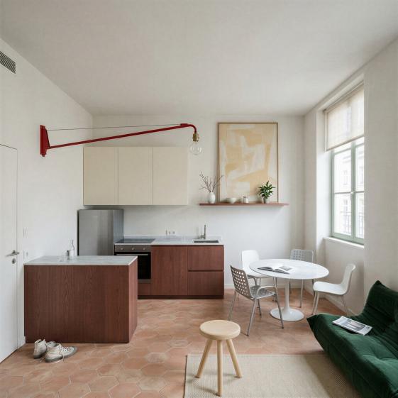 Séjour-cuisine d’un petit logement ancien : cuisine linéaire et îlot en chêne teinté brun-rouge, plan en marbre clair, sol en tomettes hexagonales, murs à l’enduit clair. Une potence rouge de Jean Prouvé au mur de gauche, un canapé Togo vert, une table ronde avec quatre chaises, un tabouret Berger au premier plan. Lumière du jour diffuse filtrée par un store en lin.