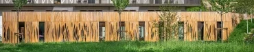 Vue extérieure de l'extension du restaurant Aribert à Saint-Martin-d'Uriage. Façade en bardage bois aux découpes organiques évoquant des flammes ou des feuilles, prairie fleurie au premier plan, terrasse et balustrade métallique en arrière-plan. Architecture biosourcée, Atelier Joëlle Personnaz. Photo D. Richalet