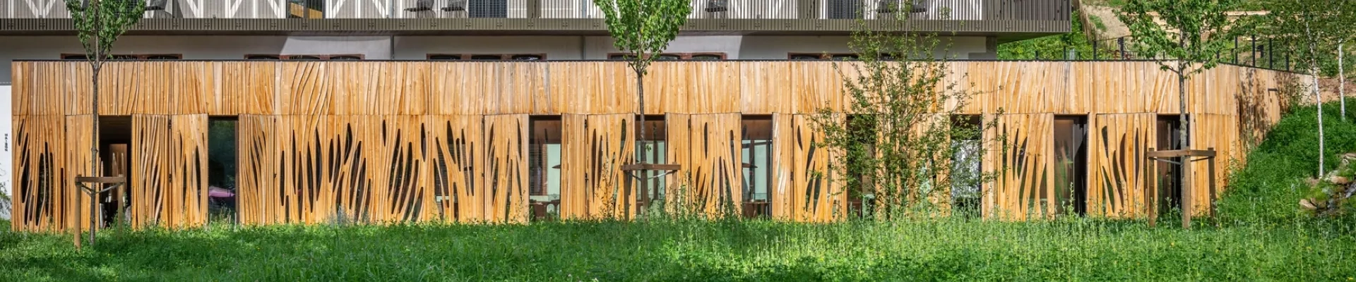 Vue extérieure de l'extension du restaurant Aribert à Saint-Martin-d'Uriage. Façade en bardage bois aux découpes organiques évoquant des flammes ou des feuilles, prairie fleurie au premier plan, terrasse et balustrade métallique en arrière-plan. Architecture biosourcée, Atelier Joëlle Personnaz. Photo D. Richalet