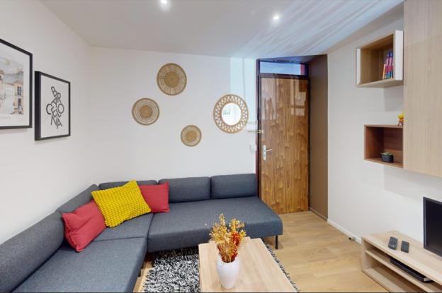 Séjour lumineux avec canapé d’angle gris, coussins colorés, table basse en bois, meuble TV et rangements muraux, dans un appartement rénové en colocation