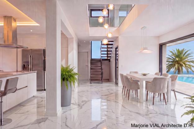 Vue du séjour de la maison contemporaine haut de gamme d'une surface plancher de 180 m². Grands volumes blancs lumineux, grandes ouvertures et lumière zénithale. Sol en marbre blanc, escalier métallique épuré, garde-corps en verre.