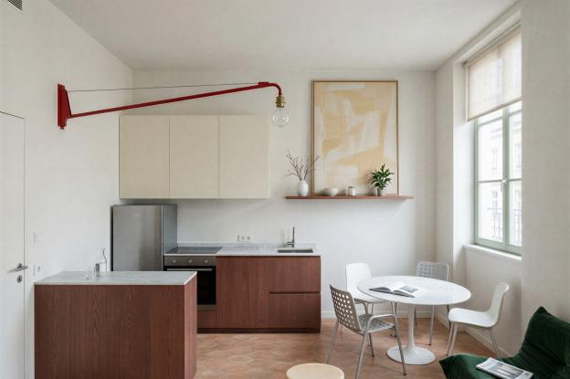 Séjour-cuisine d’un petit logement ancien : cuisine linéaire et îlot en chêne teinté brun-rouge, plan en marbre clair, sol en tomettes hexagonales, murs à l’enduit clair. Une potence rouge de Jean Prouvé au mur de gauche, un canapé Togo vert, une table ronde avec quatre chaises, un tabouret Berger au premier plan. Lumière du jour diffuse filtrée par un store en lin.